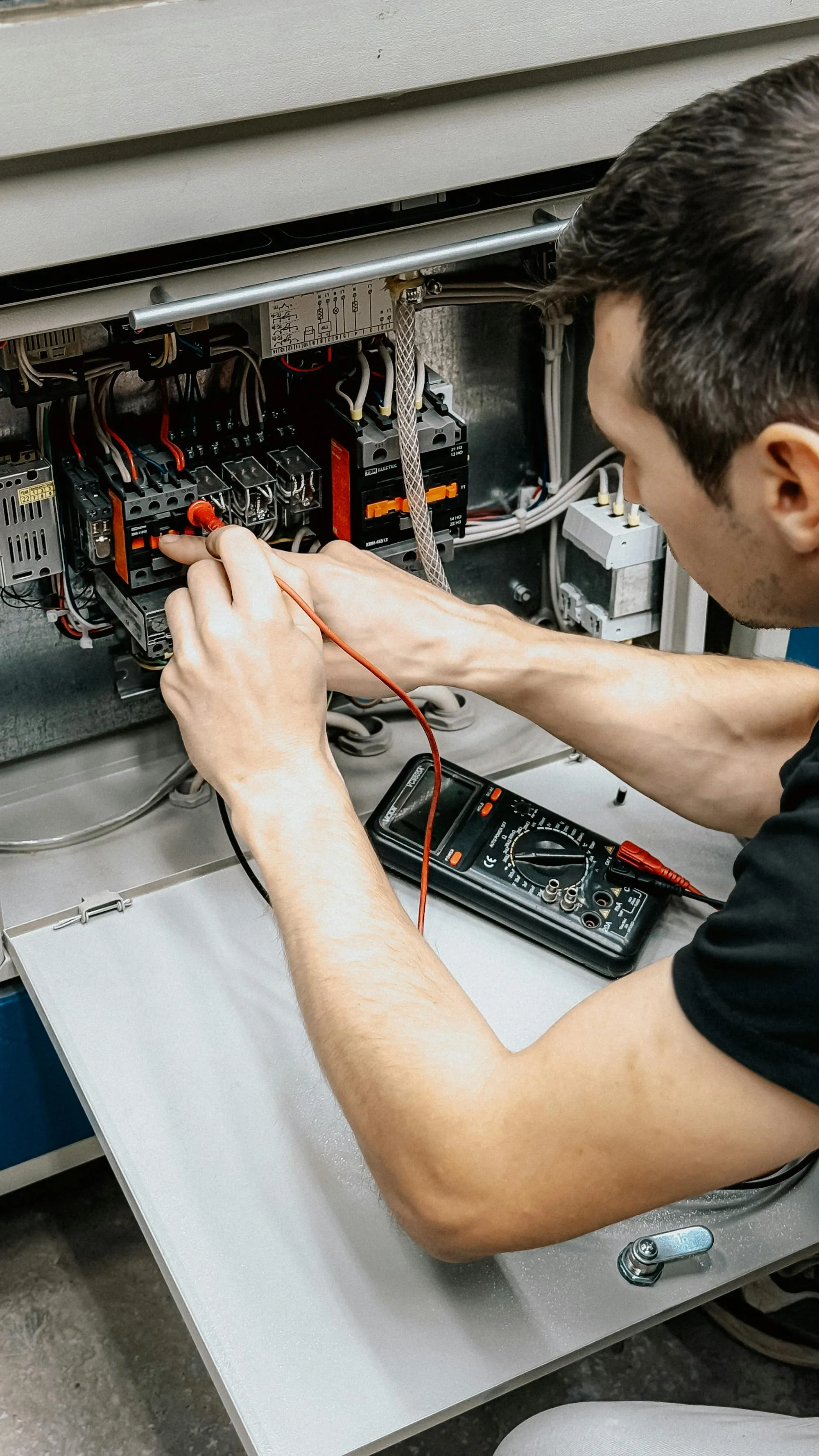 An electrician kneeling beside an open panel and testing a circuit with a multimeter.