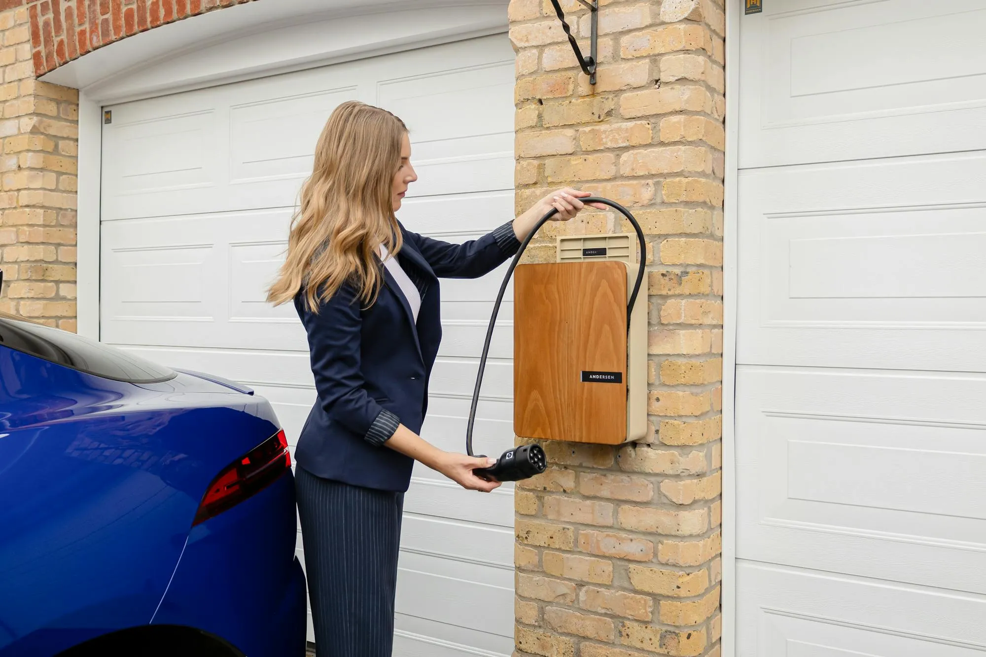 A wall-mounted home EV charger fitted to a brick pillar next to a parked electric car.